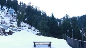 Eine hölzerne Bank in verschneiter Landschaft mit einer Steinmauer und einem Metallzaun.