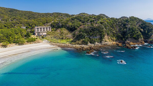 An aerial view of a coastal resort with a beachfront, a large building with multiple floors, and dense forested hills in the background. - Yado Marubun (Minamiizu)