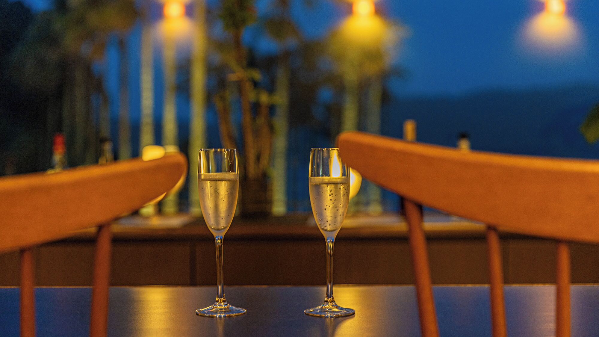 Two champagne flutes on a table with a view of the ocean at dusk.