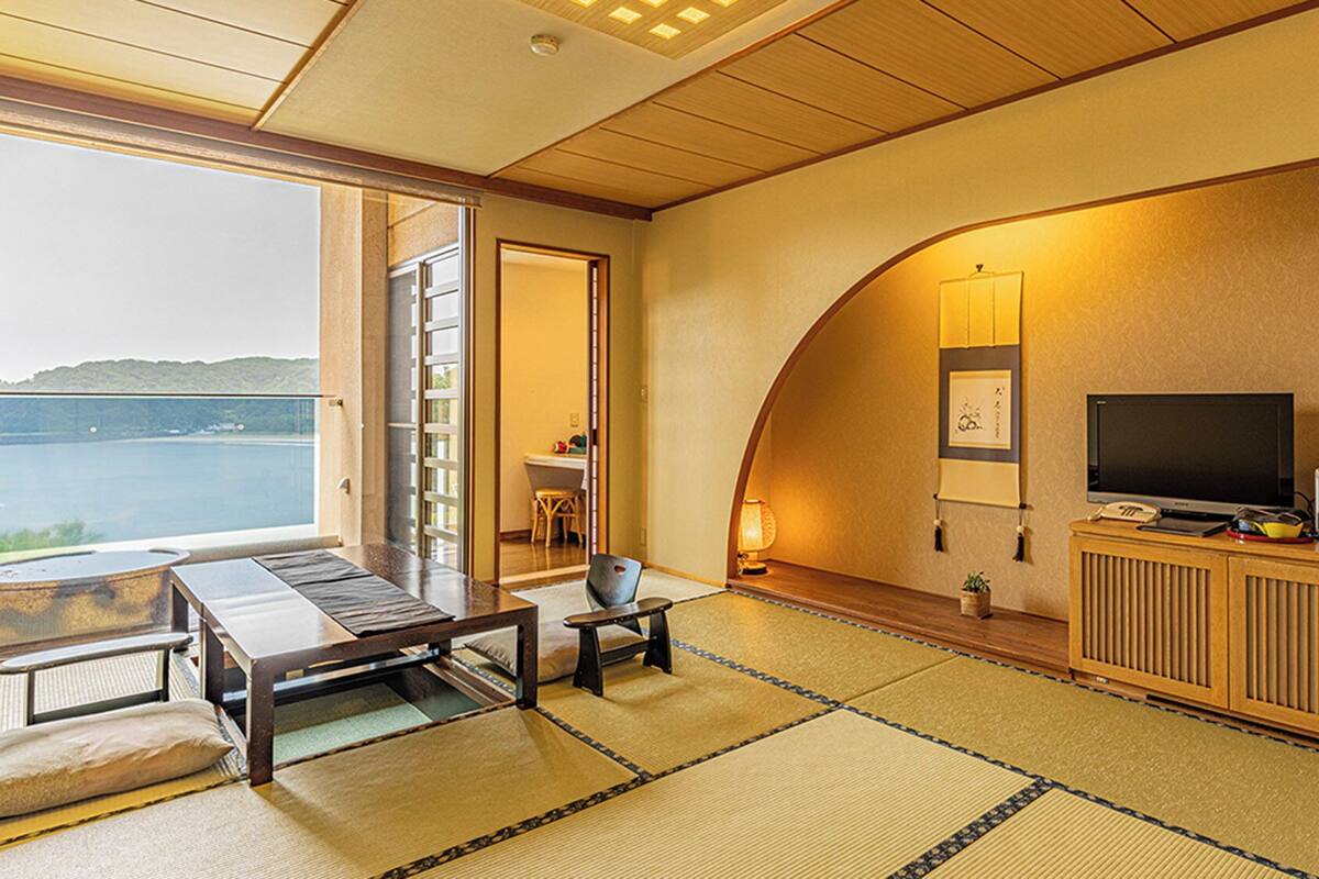 A room with tatami mats, a TV, a small table, and a view of a lake.
