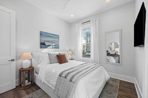 A bedroom with a bed, bedside lamps, a painting, a window with curtains, a mirror, and a door. - NEW! LSV/Golf Cart, Bikes, Walk to Beach! 2 Living Rooms & 2 Masters! Pet Friendly! (Santa Rosa Beach)