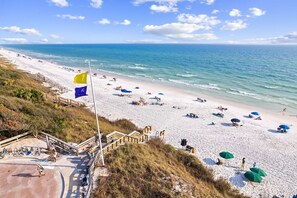 A beach with a wooden staircase leading down to the shore, a flagpole with flags, and beach umbrellas. - NEW! LSV/Golf Cart, Bikes, Walk to Beach! 2 Living Rooms & 2 Masters! Pet Friendly! (Santa Rosa Beach)