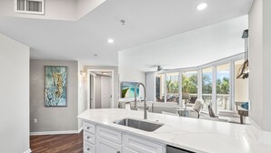 A modern kitchen with white cabinetry, a large island with a sink, and a view of the outdoors through large windows. - St Lucia 306*Guest Favorite*HighEnd*ResortAmenites (Destin)