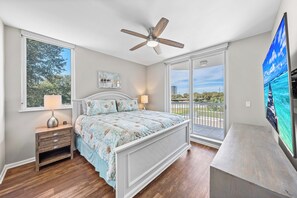 Une chambre à coucher comprenant un lit, une table de chevet, un ventilateur de plafond, un téléviseur et une grande fenêtre donnant sur l’extérieur.