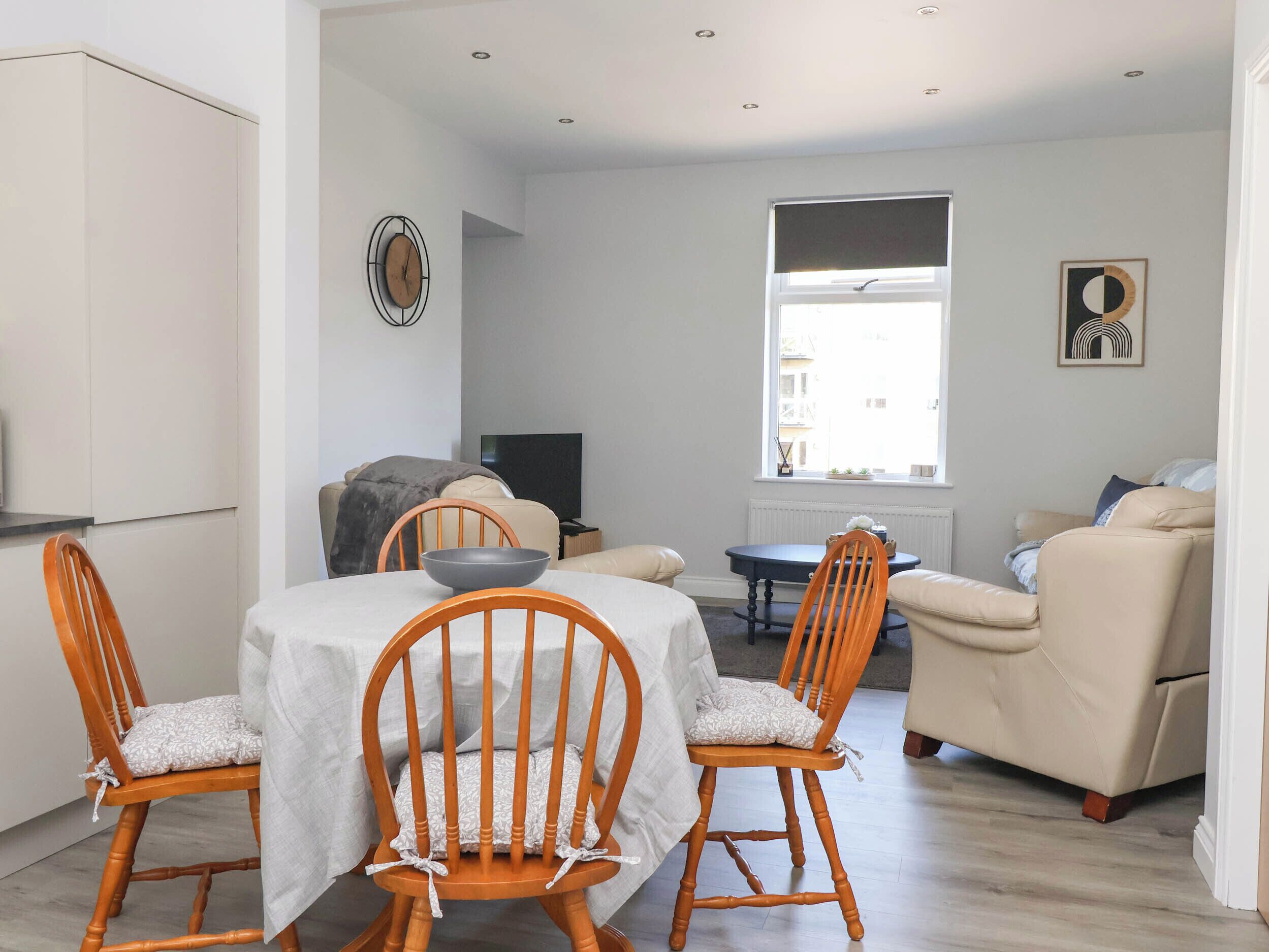 A modern living room with a dining table, two wooden chairs, a sofa, a TV, and a window with blinds.