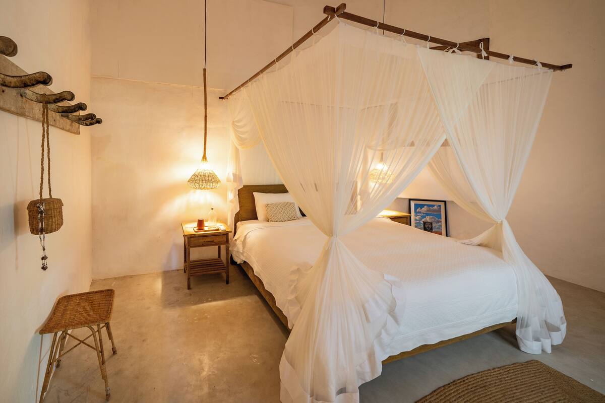 Una camera da letto con un letto a baldacchino, un comodino con una lampada, una poltrona in vimini e un cesto appeso.