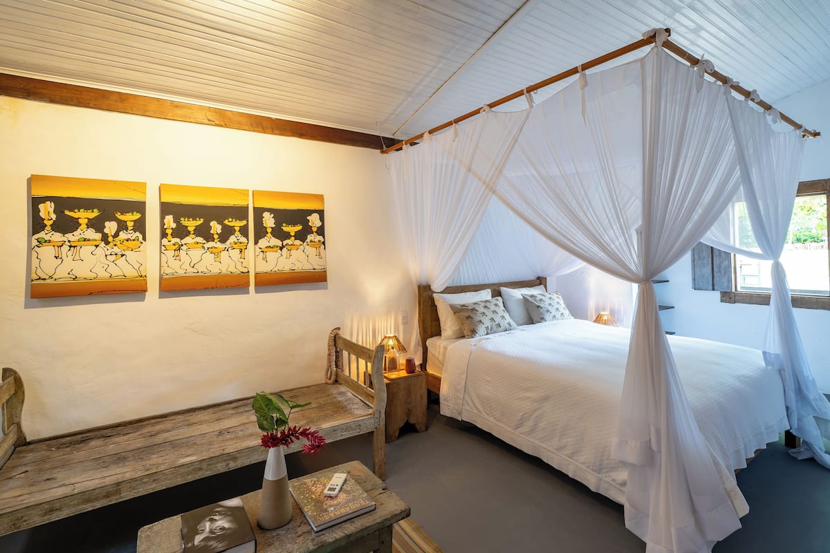 Una camera da letto con un letto a baldacchino, una panca in legno, un tavolino con un vaso e un libro, e tre quadri appesi al muro.