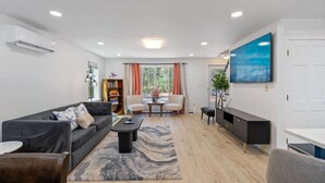 A modern living room with a sofa, coffee table, and a TV. There is a bookshelf and a dining area with a table and chairs.