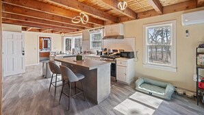 A spacious kitchen with a large island, wooden ceiling beams, and a view of trees through a window. - Cozy Riverfront Stay in Wareham (Wareham)