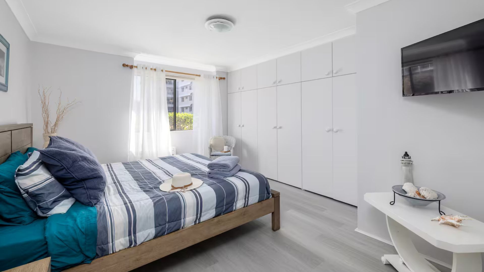 A modern bedroom with a wooden bed, a TV mounted on the wall, and a window with sheer curtains.