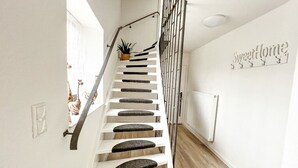 Een trap met witte leuningen en een zwarte handrail, die leidt naar een plateau met een radiator en een bordje aan de muur met daarop 'Sweet Home'.