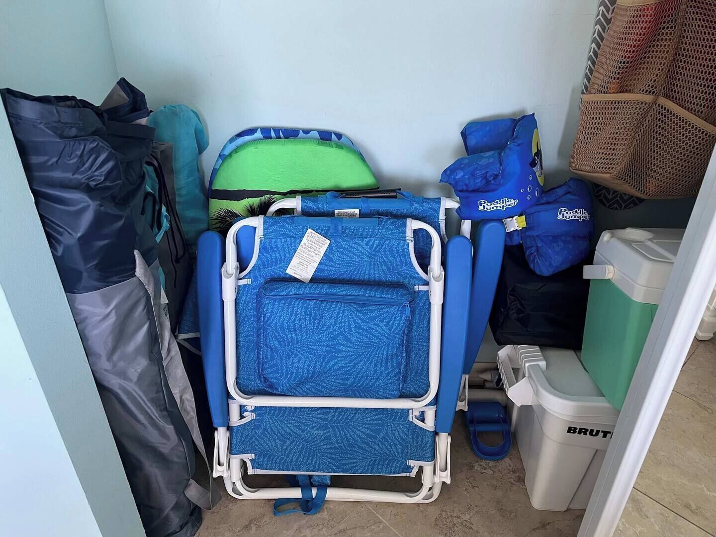 A small, cluttered room with a blue portable bed, a green suitcase, and a blue bag labeled 'Puddle Jumper'.
