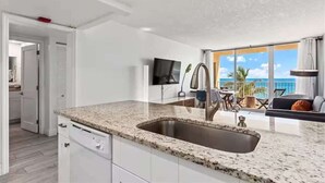 A modern kitchen with a granite countertop and stainless steel sink, a dining area with a view of the ocean, and a flat-screen TV mounted on the wall.