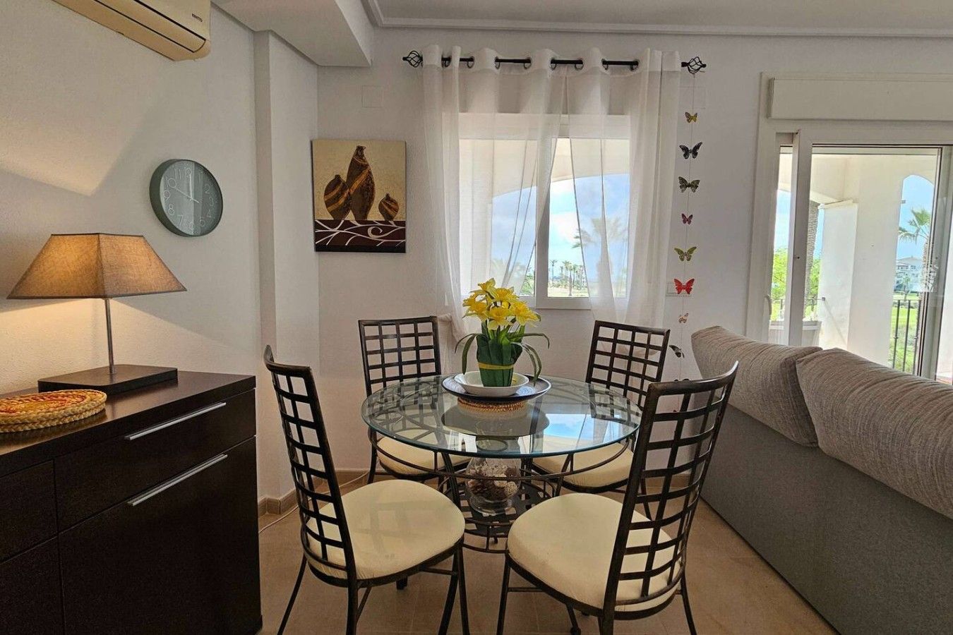 Un coin repas comprenant une table ronde en verre, quatre chaises, une lampe, une horloge, un tableau et un canapé.
