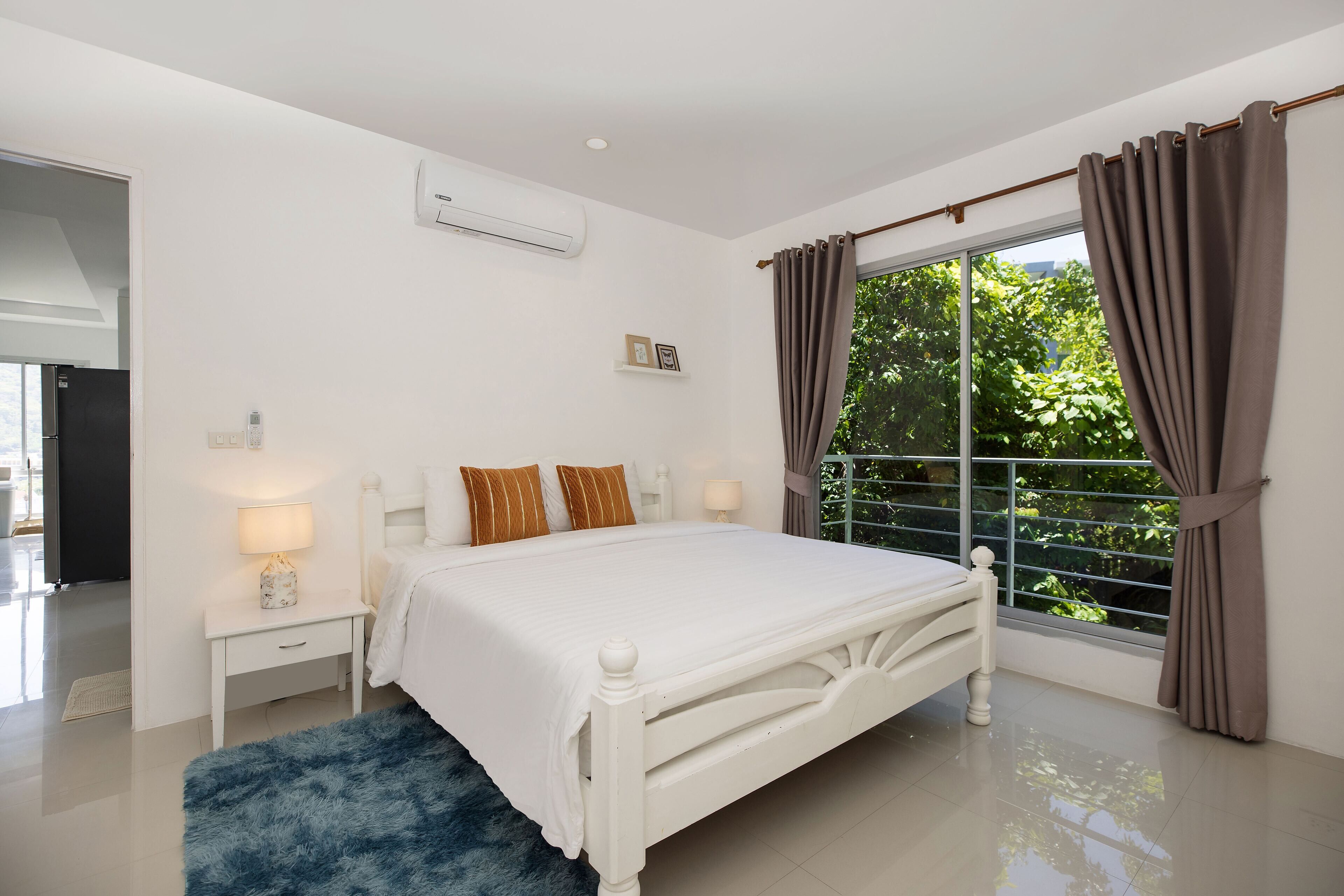 A bedroom with a white bed, a nightstand with a lamp, a window with curtains, and a view of greenery outside.