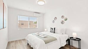A modern bedroom with a large bed, a bedside table with a lamp, and a window with blinds. - Modern Home Near Pasadena Garage Full Kitchen (Alhambra)