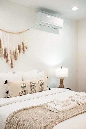 Un lit bien fait, avec du linge de lit blanc et un plaid beige, une lampe de chevet et un climatiseur fixé au mur.
