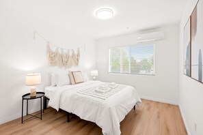 Une chambre à coucher au style minimaliste, avec un grand lit, des tables de chevet, un climatiseur fixé au mur et une fenêtre équipée de stores.