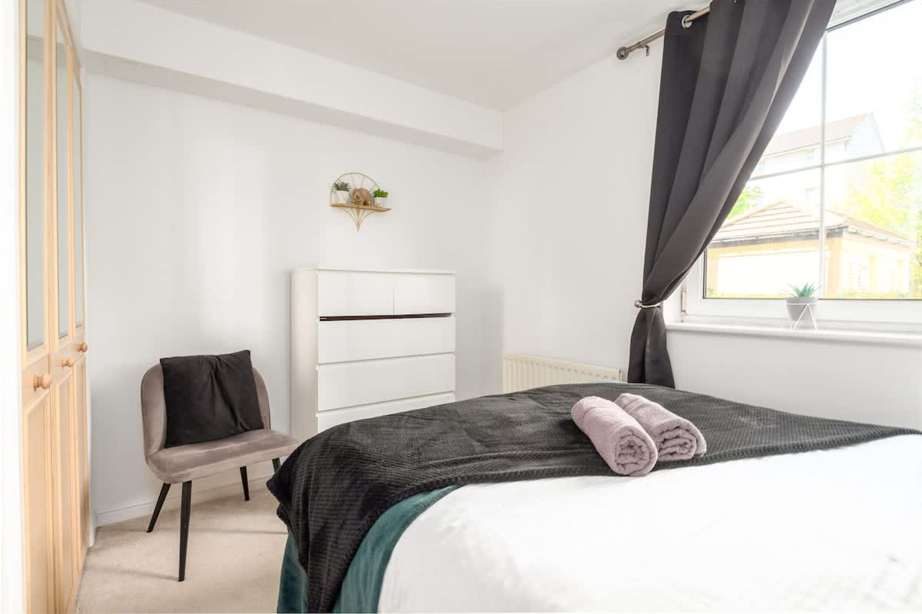 Un dormitorio con cama, una silla, una estantería con plantas y una ventana con cortinas.
