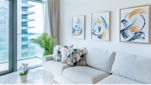 A modern living room with a sofa, abstract art, a potted plant, and a view of a high-rise building. - Homesgetaway-luxury 2Br Apartment in Beach Vista Tower 2 With Beach Access (Dubai)
