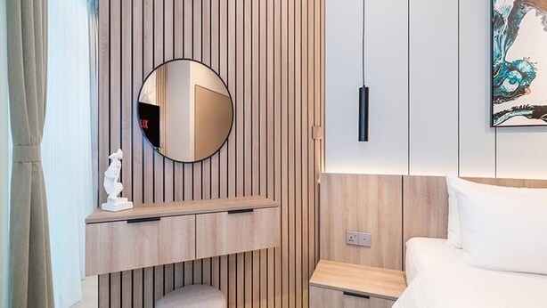 Moderni hotellihuone, jossa on puupanelointi yhdellä seinällä, pyöreä peili, yöpöytä, pieni jakkara ja valkoisilla vuodevaatteilla varustettu sänky.
