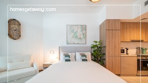 A modern hotel room with a bed, a small bedside table, a nightstand, a wall-mounted artwork, a potted plant, and a kitchenette with wooden cabinets. - Sophisticated Studio Escape Missoni Luxury Design (Dubai)