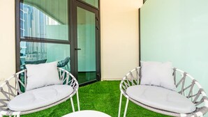 Two wicker chairs with white cushions on a green artificial turf, positioned on a balcony with a glass door and a view of tall buildings. - Sophisticated Studio Escape Missoni Luxury Design (Dubai)