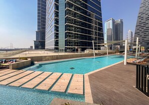Une piscine sur le toit, dotée d’une terrasse en bois avec des chaises longues, offrant une vue sur les gratte-ciel et un ciel dégagé.