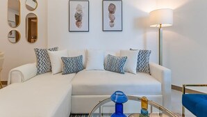 Una sala de estar moderna con un sofá blanco, cojines estampados, una mesa de centro de vidrio y obras de arte enmarcadas en la pared.