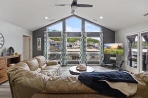 A spacious room with a large window, a ceiling fan, a sofa, a coffee table, and a television. - Beautiful Lake Front Home with Private Dock In Lick Branch Cove (Rocky Mount)