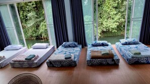 Una habitación con cuatro camas, cada una con una colcha y almohadas estampadas, un ventanal con cortinas y un ventilador de pie.