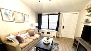 A living room with a beige sofa, a coffee table, a TV stand, and a window with blinds. - Charming  1 bedroom apartment with AC, WiFi in quaint Columbus (Columbus)