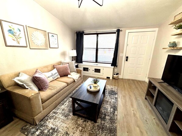 A living room with a beige sofa, a coffee table, a TV stand, and a window with blinds.