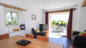 A modern living room with a wooden dining table, a sofa, and a TV. There is a large window and a sliding glass door leading to an outdoor patio with a table and chairs. - Great Hartbarrow Farm Cottage (Windermere)