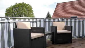 Two wicker chairs with beige cushions on a wooden deck, a metal railing, and a red-tiled roof in the background. - Minihotel (Pforzheim)