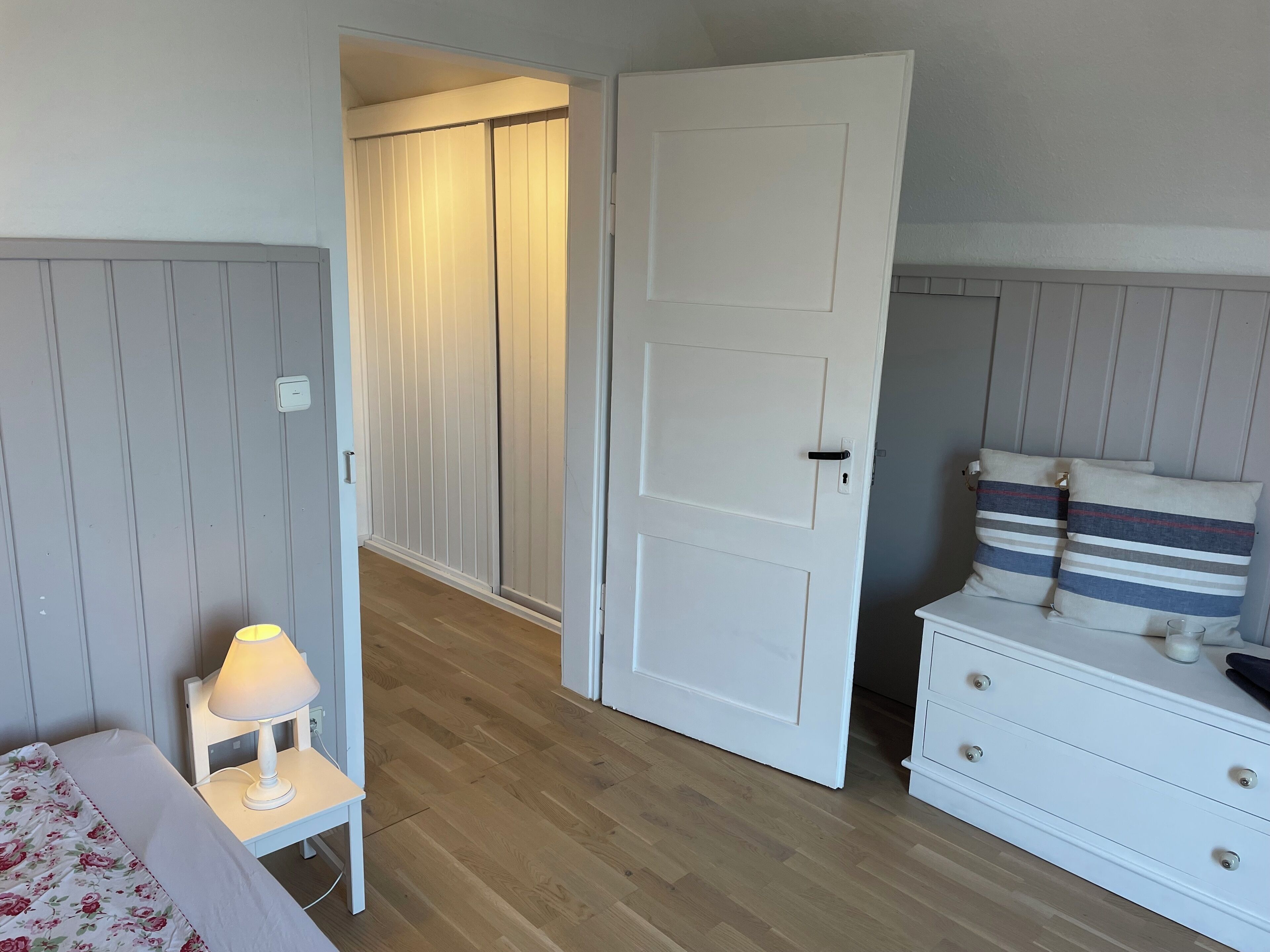 A bedroom with a bed, bedside table, lamp, wardrobe, and a dresser with striped pillows.