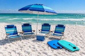 A beach scene with four striped beach chairs, a blue and white beach umbrella, a cooler, and a surfboard.