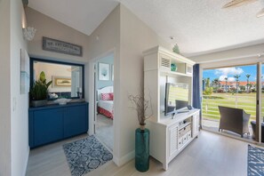 A modern living room with a TV, a blue cabinet, a dining area with a table and chairs, and a view of a garden and palm trees.