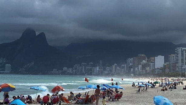 Una playa concurrida con sombrillas y gente, un horizonte urbano al fondo y montañas.