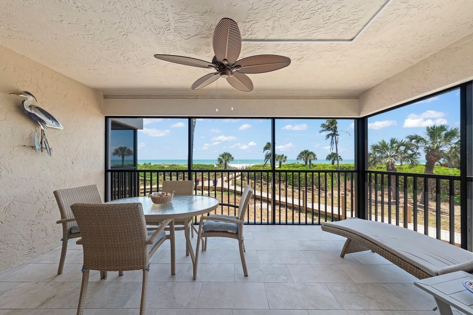 A balcony with a table and chairs, a ceiling fan, and a view of the beach and palm trees.
