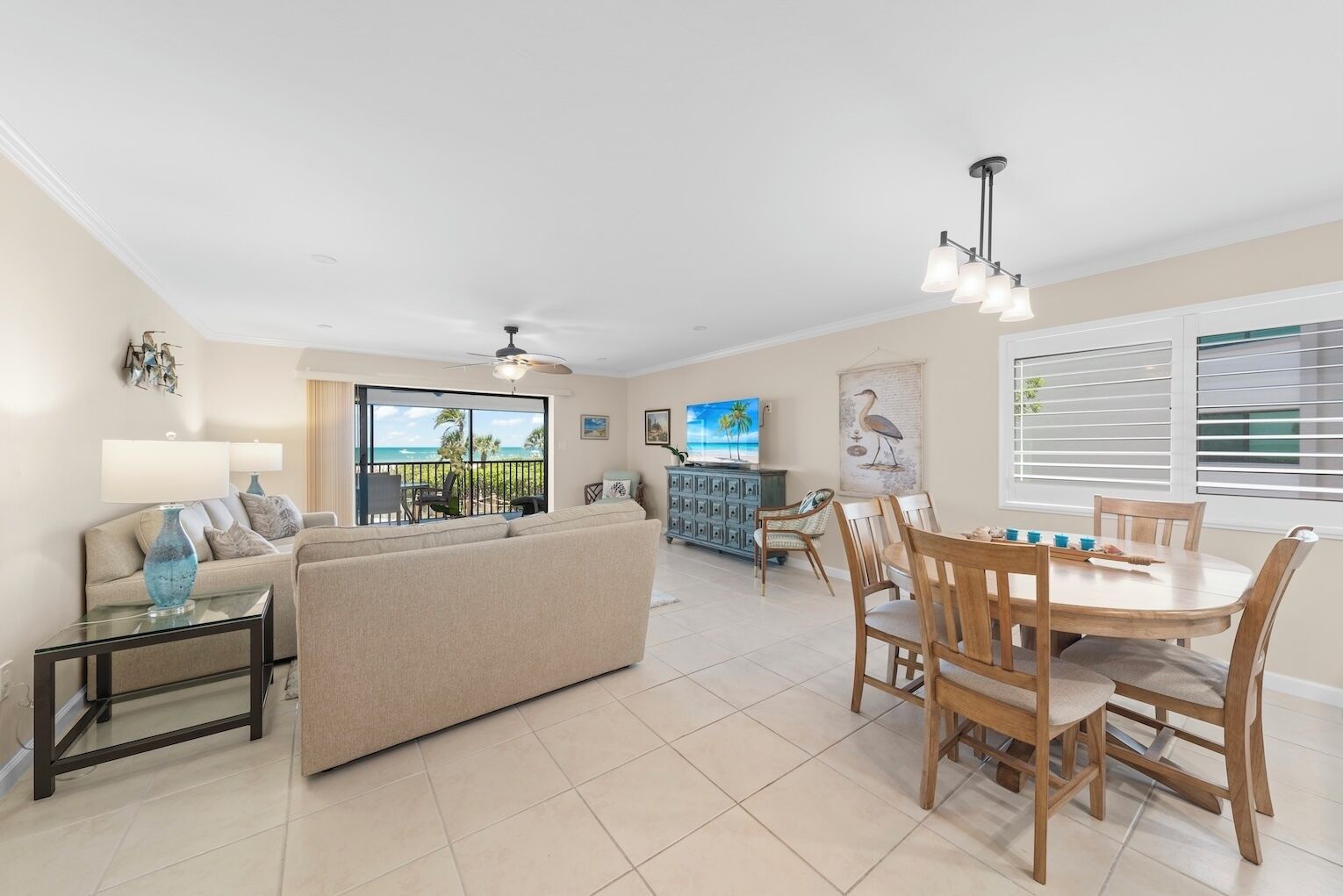 A spacious living room with a sofa, a dining table with chairs, a TV, and a balcony with a view of the ocean.