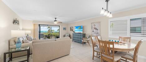 A spacious living room with a sofa, a dining table with chairs, a TV, and a balcony with a view of the ocean.