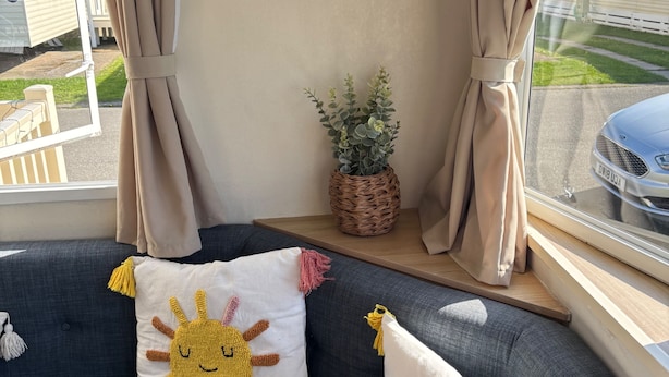 Una sala de estar acogedora con un sofá gris, dos cojines con motivos de sol, una frase inspiradora en un marco, una planta en maceta y vistas a una zona residencial.
