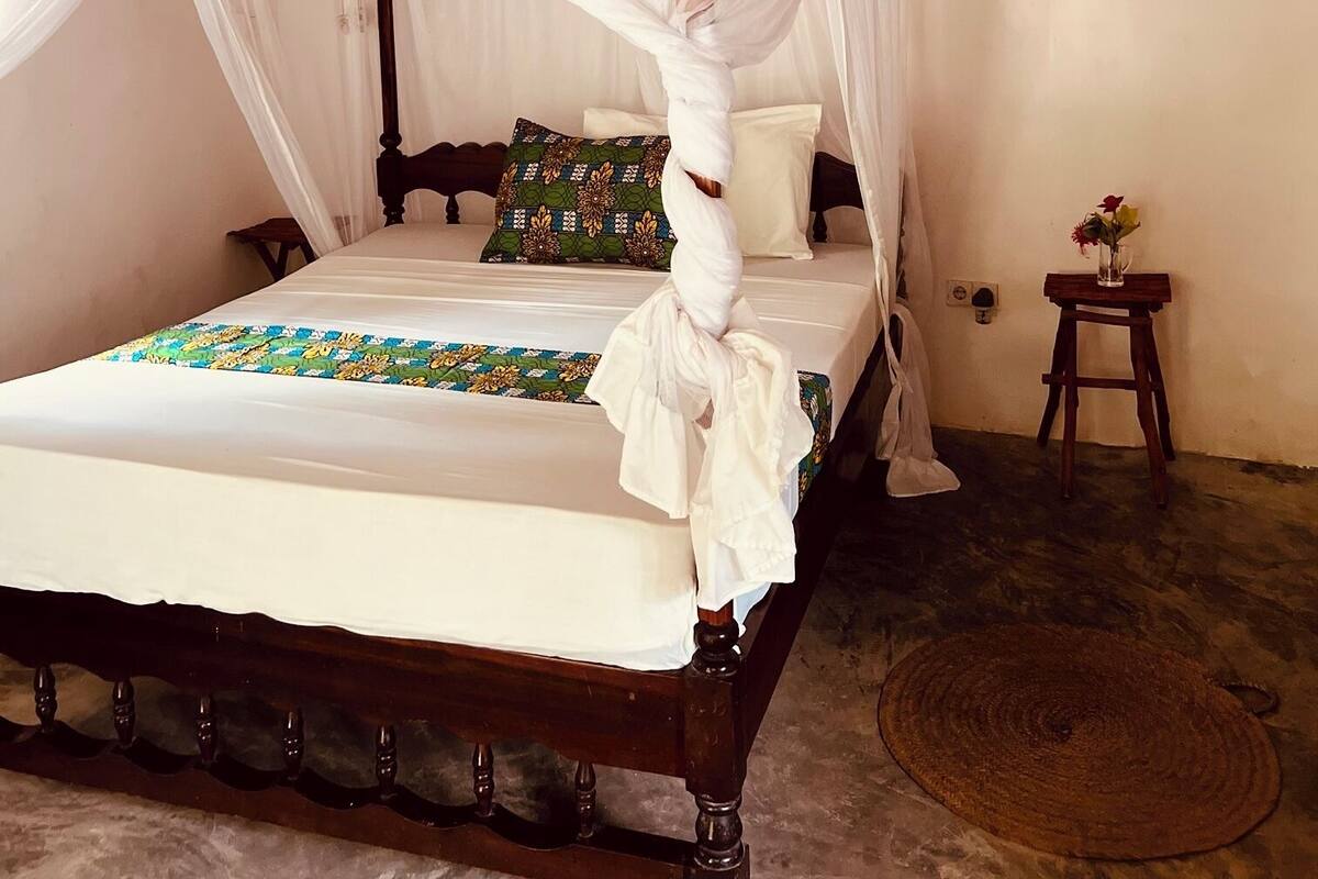 A bed with a canopy, a wooden headboard, a patterned bedspread, and a small bedside table with a vase.