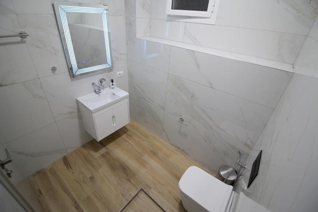 Une salle de bain avec un lavabo, un miroir et des toilettes blancs, un plancher en bois et des murs en marbre.
