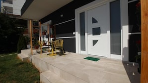 A modern building with a black exterior and a white door. There are steps leading to a covered porch area with a yellow chair and potted plants. - Alis Apartments (Sarande)