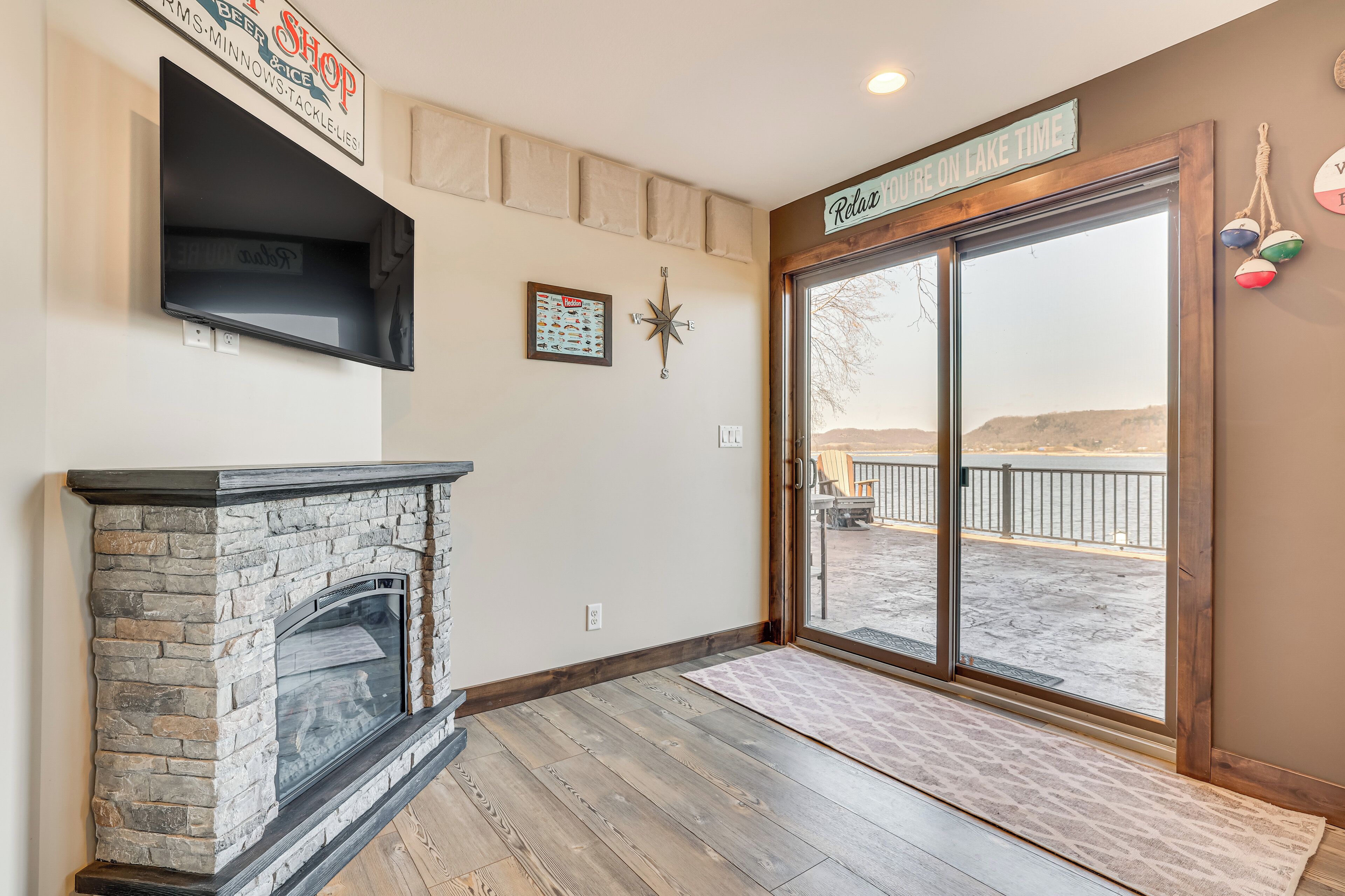 A room with a stone fireplace, a large window with a view of a lake, and a sign that reads "Relax on Lake Time."