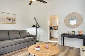 Una sala de estar moderna con un sofá gris, una mesa de centro redonda de madera, una lámpara de pie negra y una cocina visible a través de un vano abierto.