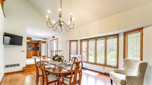 Una sala da pranzo con un tavolo in legno, sedie e un lampadario. Si intravede una cucina sullo sfondo, con pensili in legno e un frigorifero.