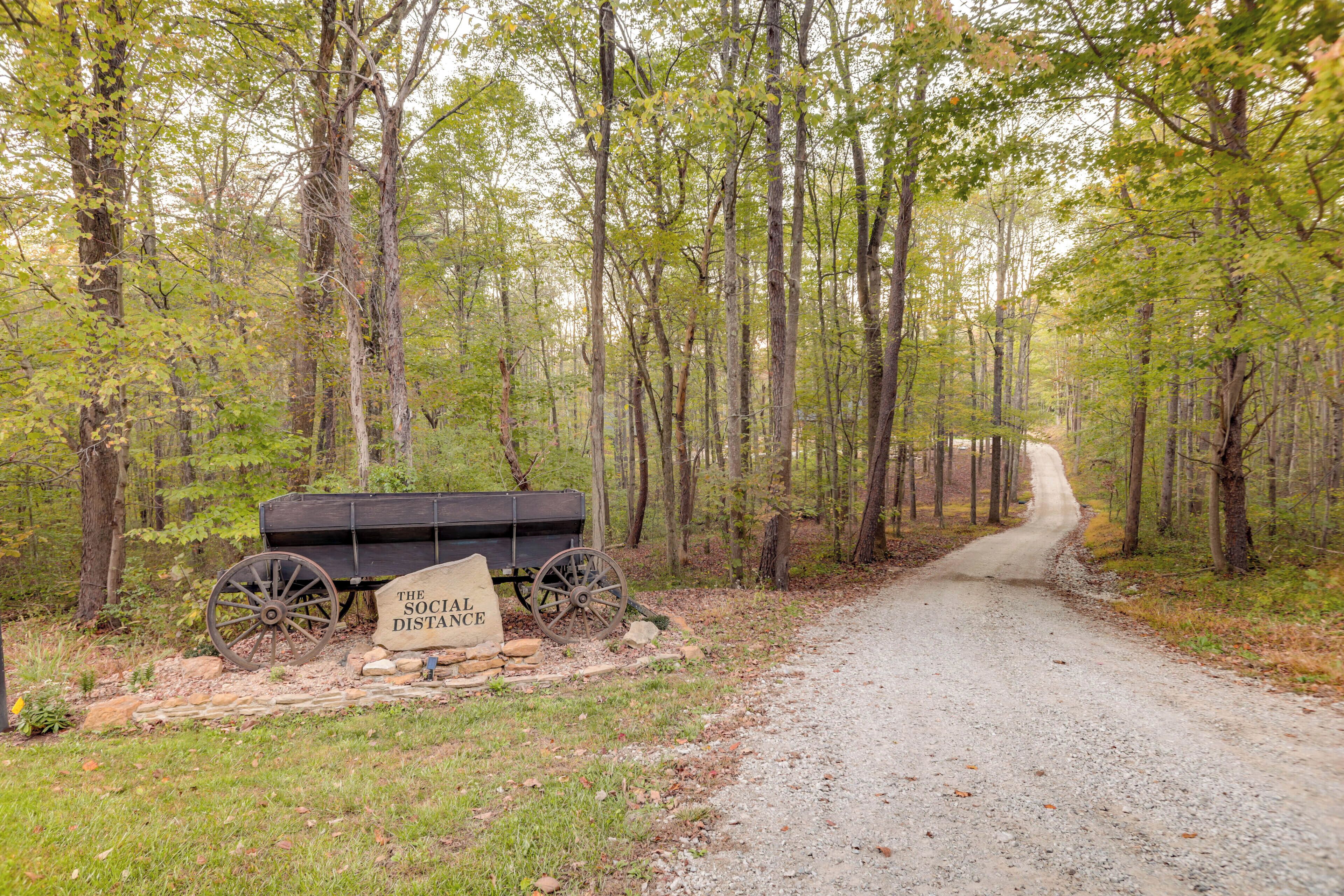 A gravel path leading through a forest with a wooden cart labeled "The Social Distance" and a sign.